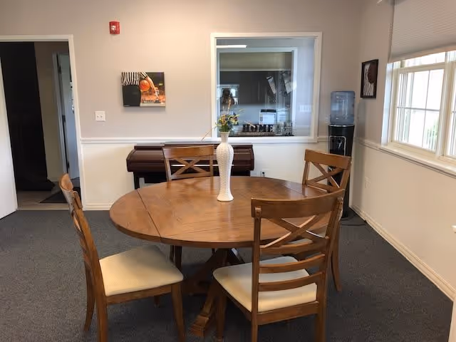 Interior room with a round wooden dining table surrounded by four wooden chairs with beige cushions. A white vase with flowers is placed in the center of the table. In the background, there is a piano against the wall, a window with a view into another room, a water dispenser, and a window letting in natural light.