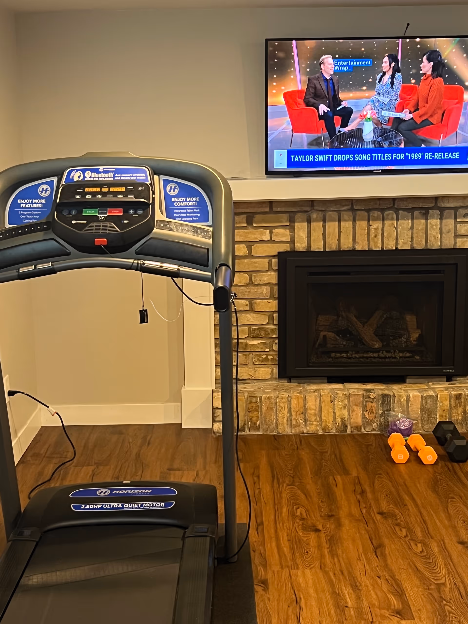 Treadmill in a living area with fireplace, mounted TV, and dumbbells nearby.