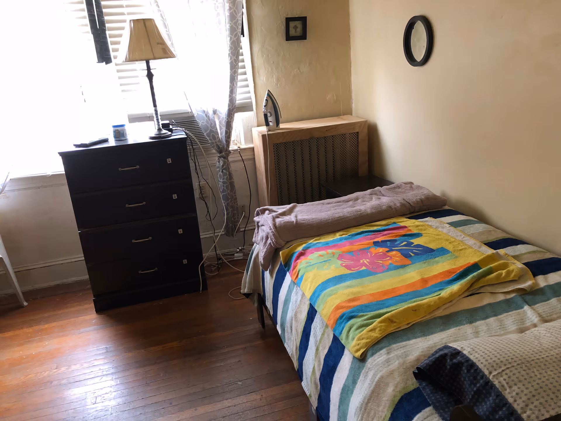 A small bedroom with a bed, dresser, lamp, and window blinds.