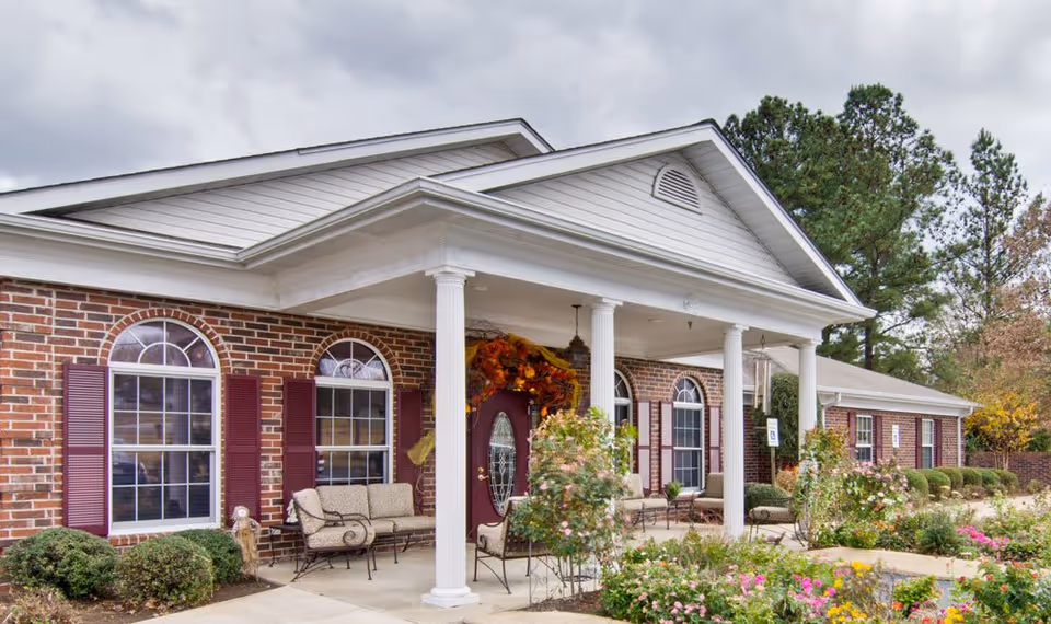 Exterior view of a brick building with white columns supporting a covered entrance. The building has arched windows with maroon shutters and a maroon door decorated with a fall-themed wreath. There are benches on the porch and landscaped bushes and flowers in front of the building under a cloudy sky.
