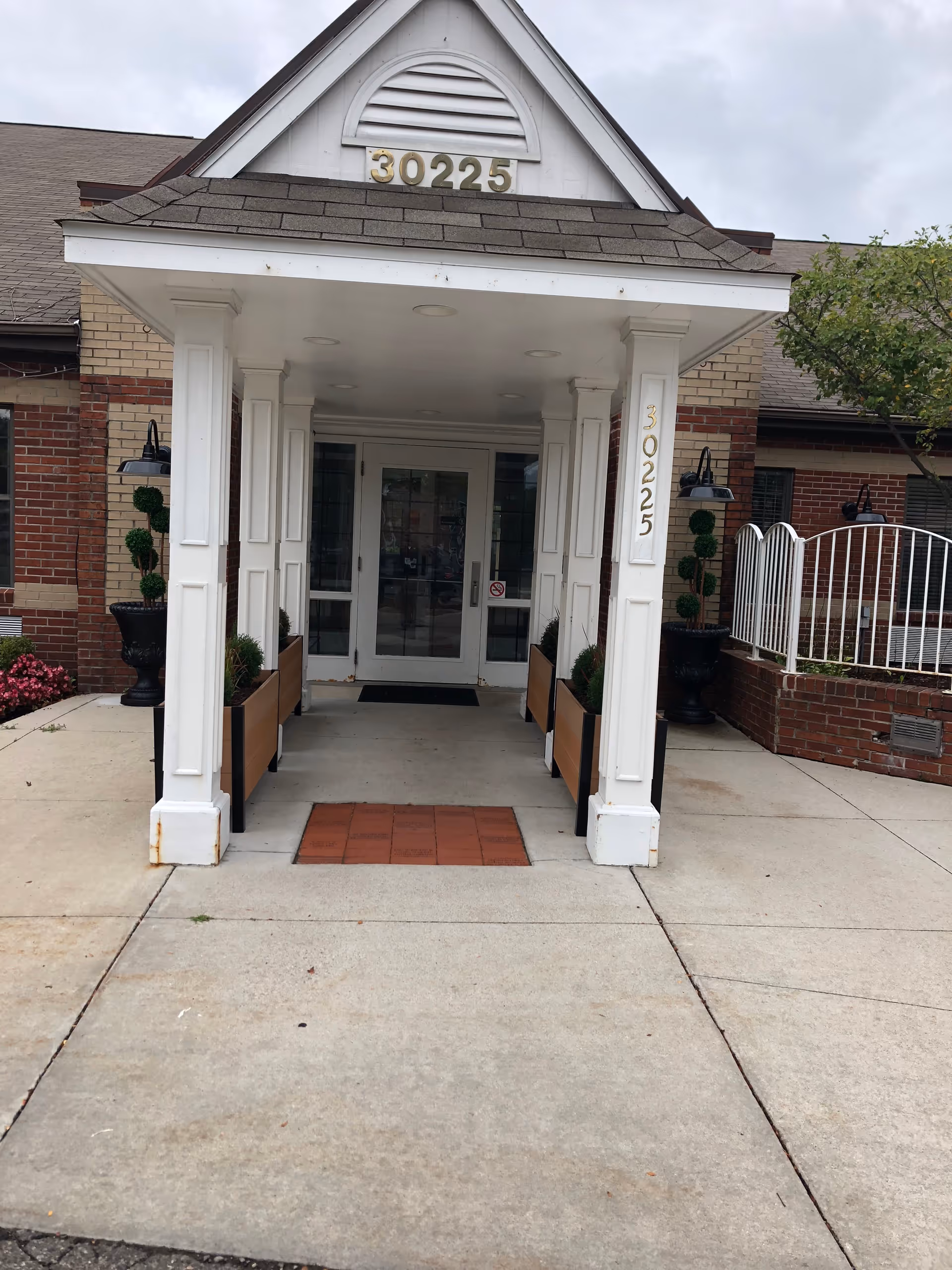 Entrance to a building with a covered porch supported by white columns. The building has brick walls and the number 30225 is displayed above the entrance and on one of the columns. There are potted plants on either side of the entrance and a small tree to the right.