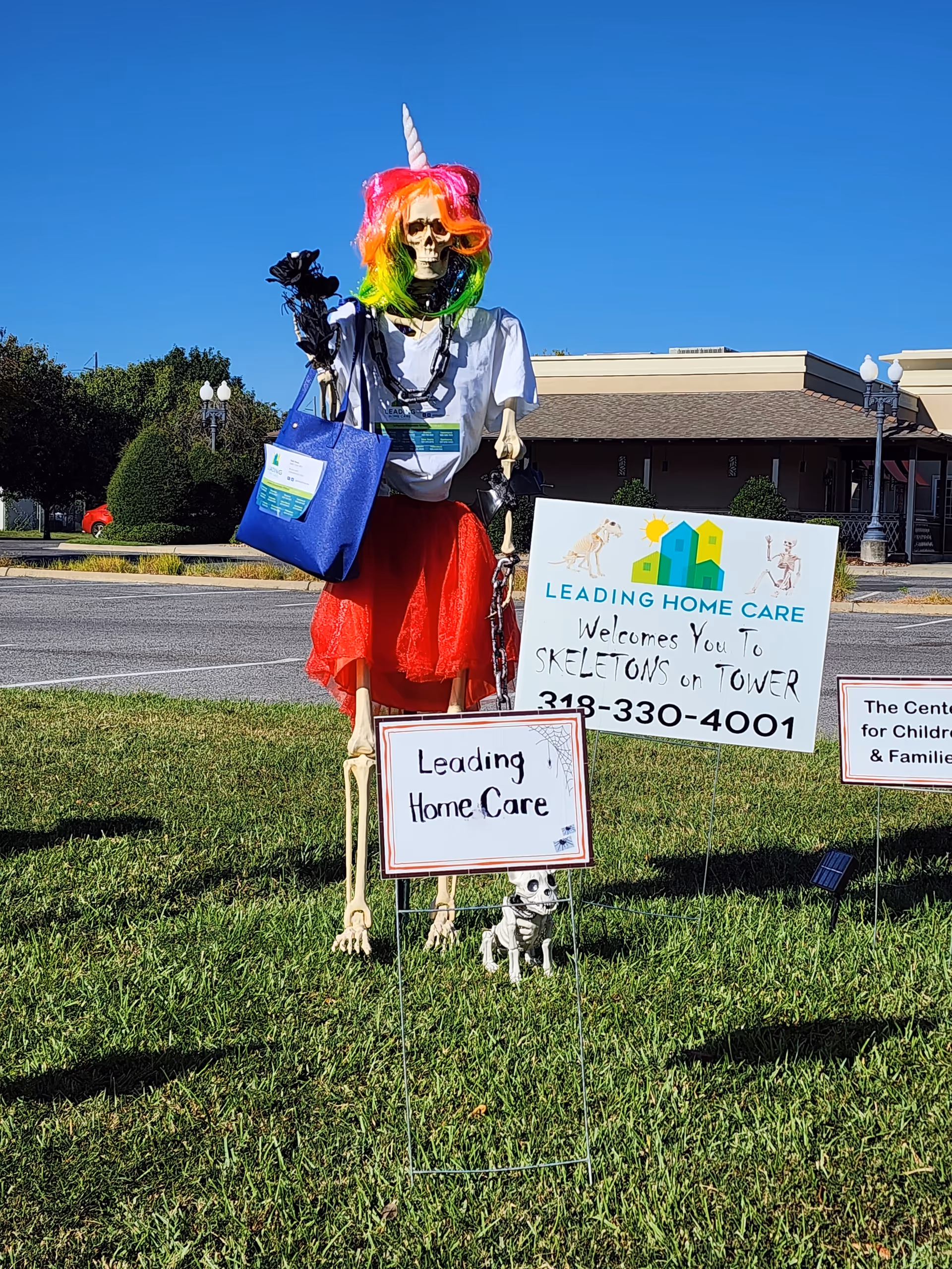 Skeleton decoration with rainbow wig and tote bag beside Leading Home Care signs outside building.