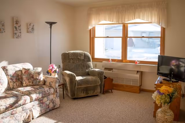 A cozy living room with a floral patterned sofa, a green recliner chair, a floor lamp, a small side table with a flower vase, a window with sheer curtains letting in sunlight, and a TV on a wooden stand with a flower arrangement beside it.