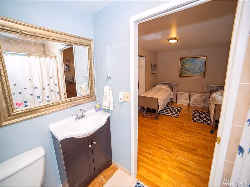 View from a bathroom into a bedroom. The bathroom has a white sink with a dark brown cabinet, a large ornate mirror, a towel hanging on the wall, and a toilet partially visible. The bedroom has two single beds with light-colored bedding, two small white dressers between the beds, a wooden floor, and a framed painting on the wall.