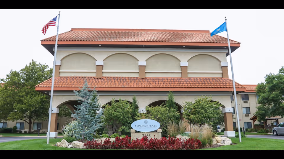Front entrance of the Sedgwick Plaza building with flags, arched portico, landscaped beds, and a sign.