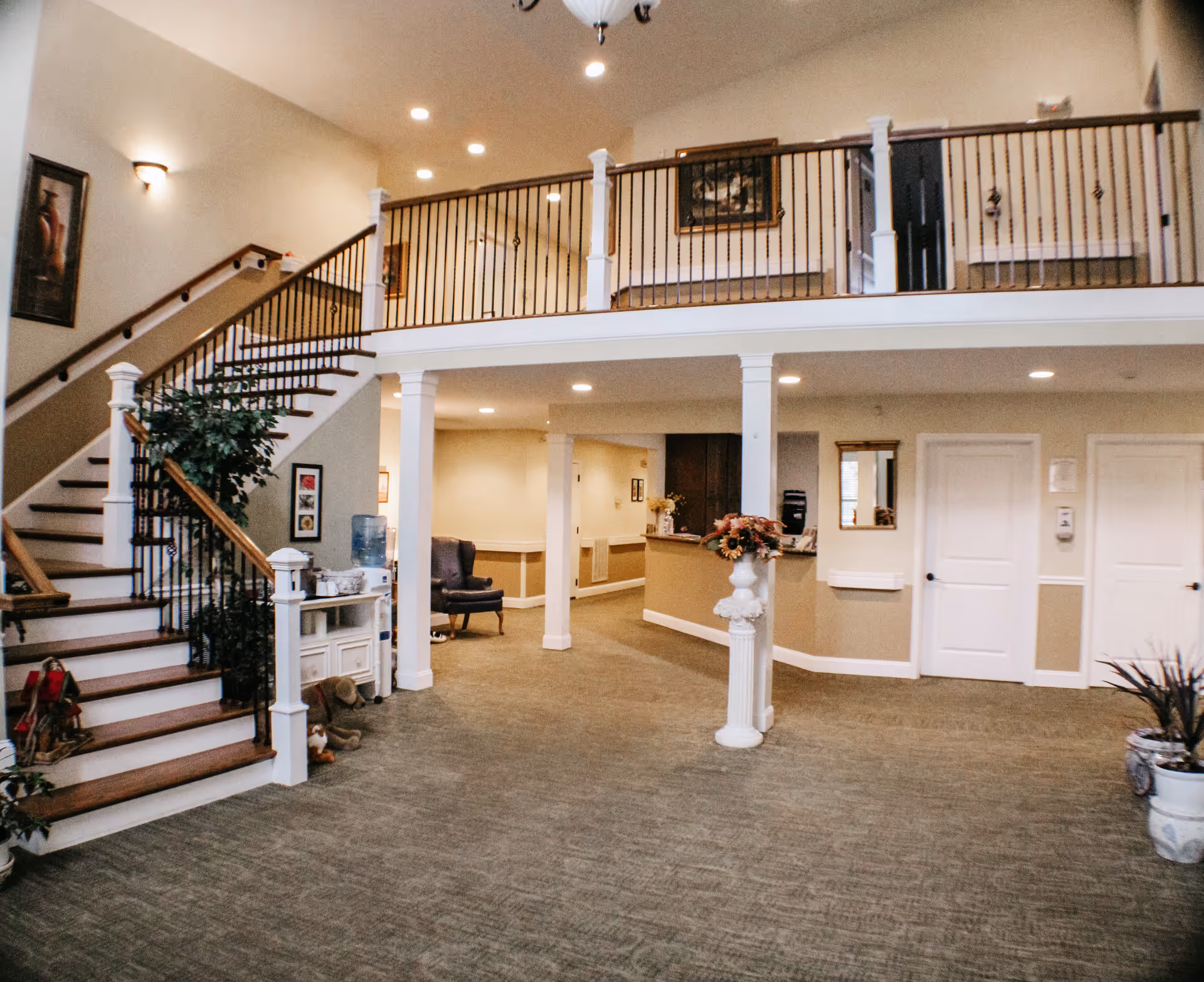 Interior view of a senior living facility lobby area with a staircase on the left, a water cooler, a chair, and a reception desk in the background. The space has beige walls, carpeted floors, and decorative plants and artwork.