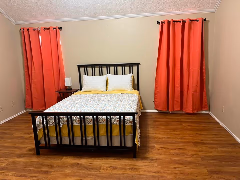 A black metal bed frame with yellow bedding in a simple bedroom with red curtains.