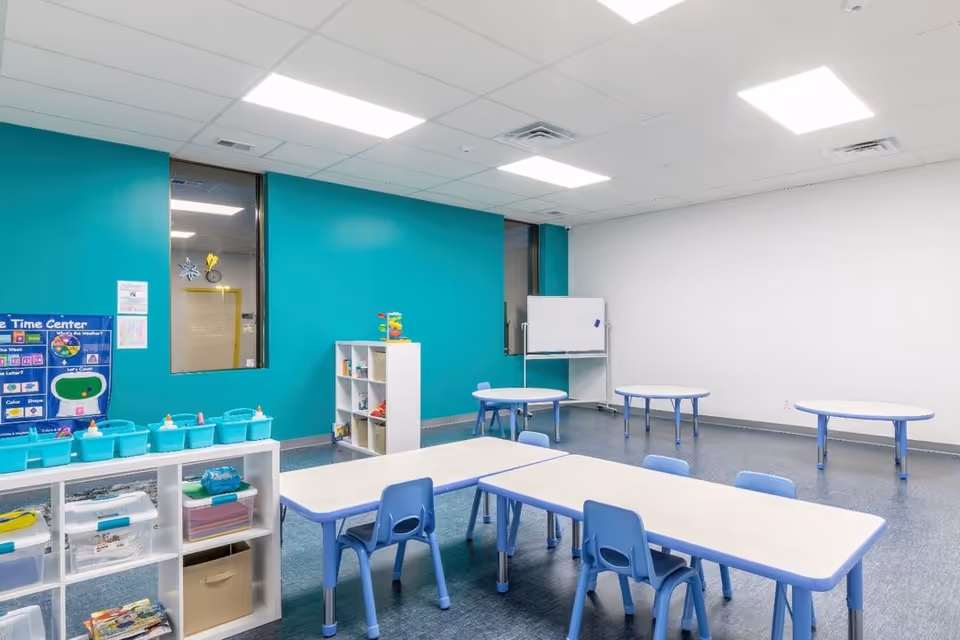 Bright teal-walled classroom with small blue tables, chairs, and toy storage shelves.