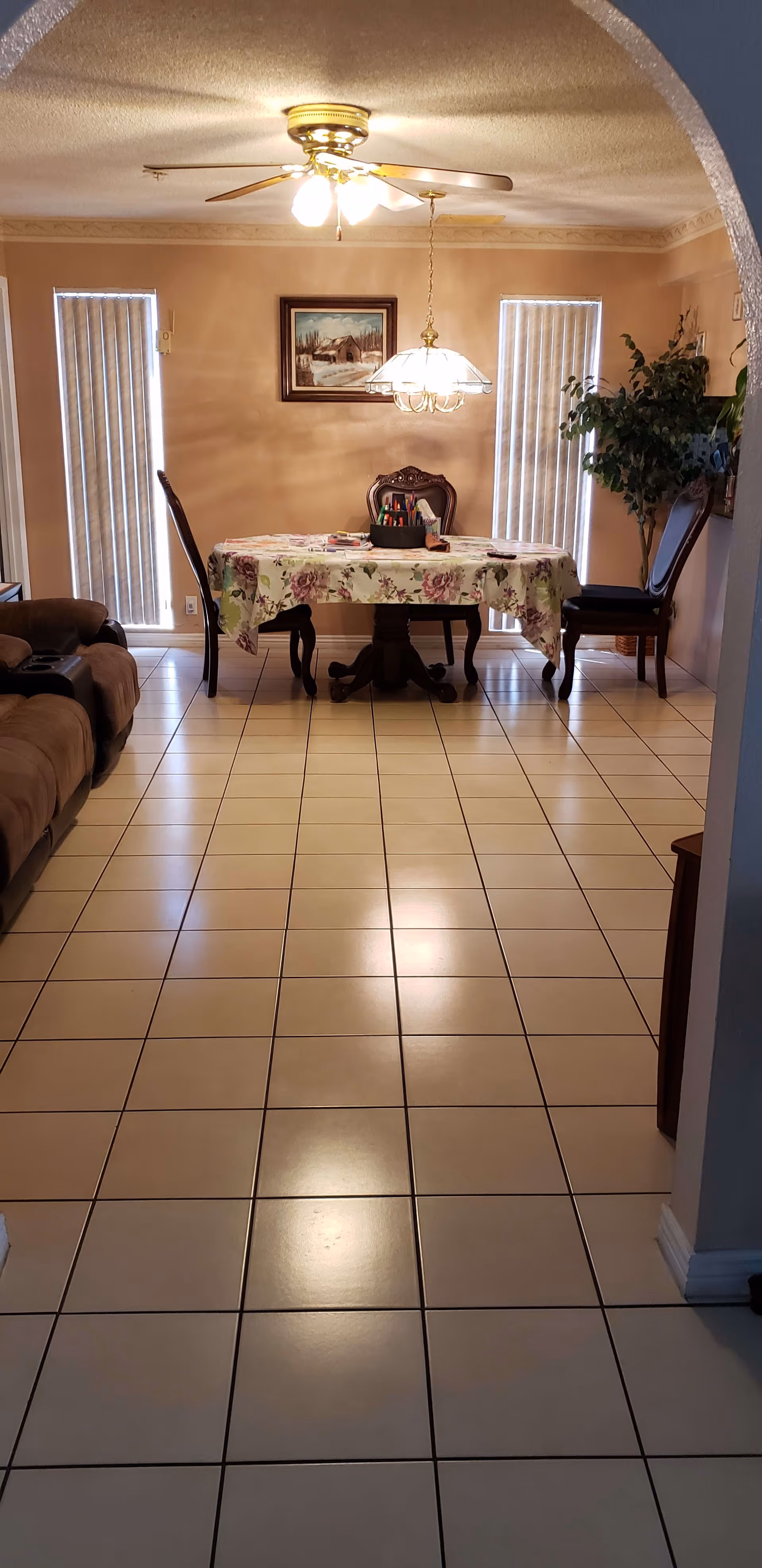 Interior view of a dining area with a rectangular table covered with a floral tablecloth, surrounded by four wooden chairs. The room has tiled flooring, two vertical blinds covering windows on the back wall, a ceiling fan with lights, a hanging light fixture above the table, a framed painting on the wall, a potted plant in the corner, and a brown sofa partially visible on the left side.