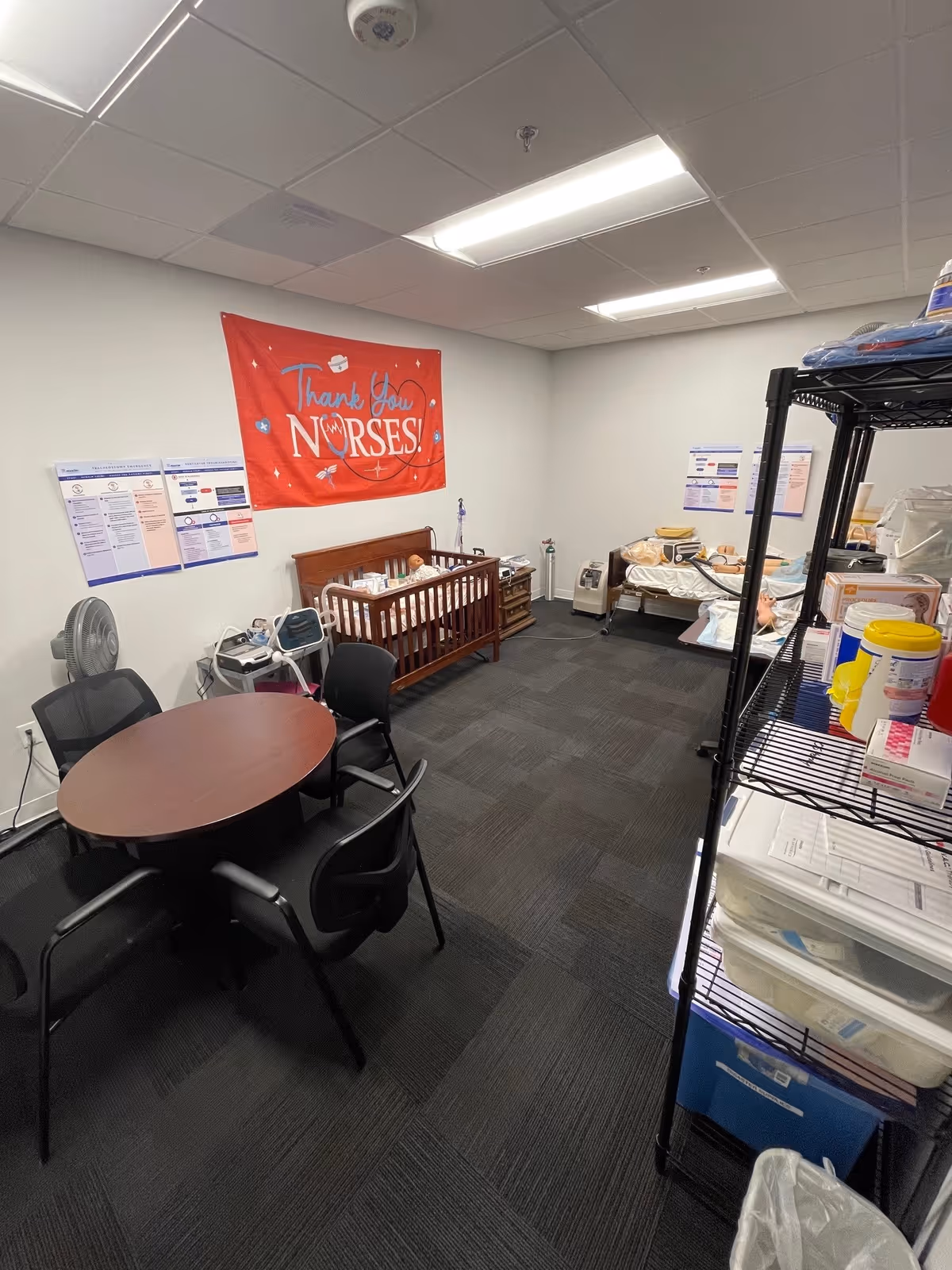 Interior medical room with round table, chairs, crib, medical supplies, and shelving.
