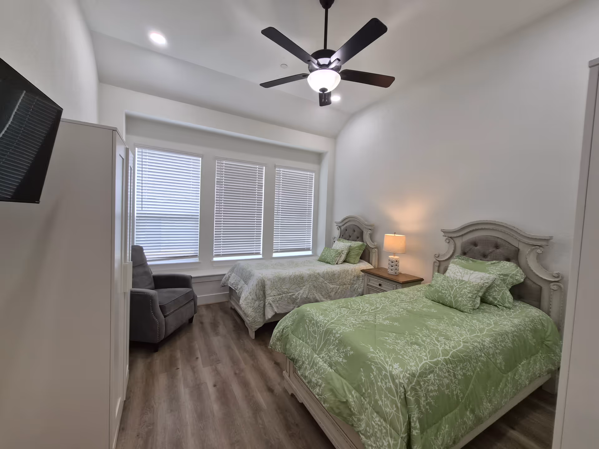 Two twin beds with green bedding and a nightstand lamp in a bright bedroom.
