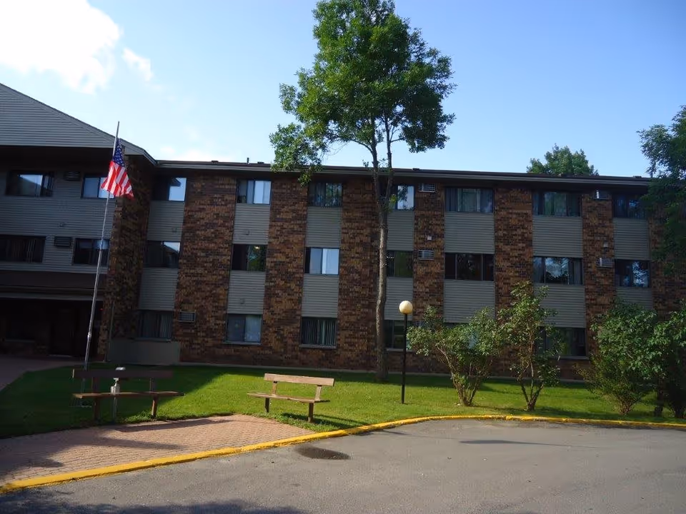 Front view of a three-story brick-and-siding senior living building with an American flag, benches, a lamppost, and trees on a grassy courtyard.
