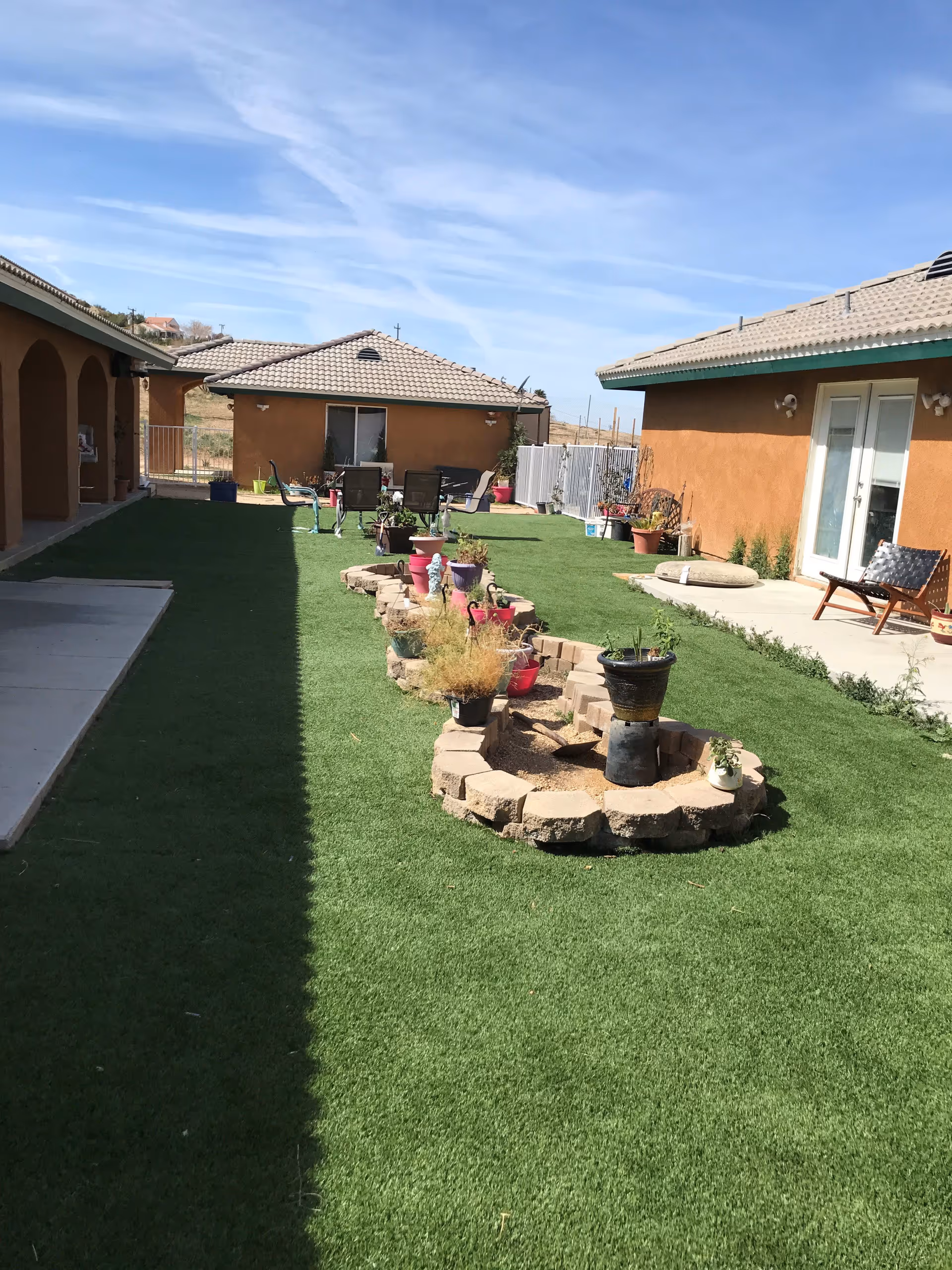 Outdoor courtyard area with artificial grass, a central raised garden bed made of bricks containing various potted plants, surrounded by single-story buildings with tan stucco walls and tiled roofs under a blue sky.
