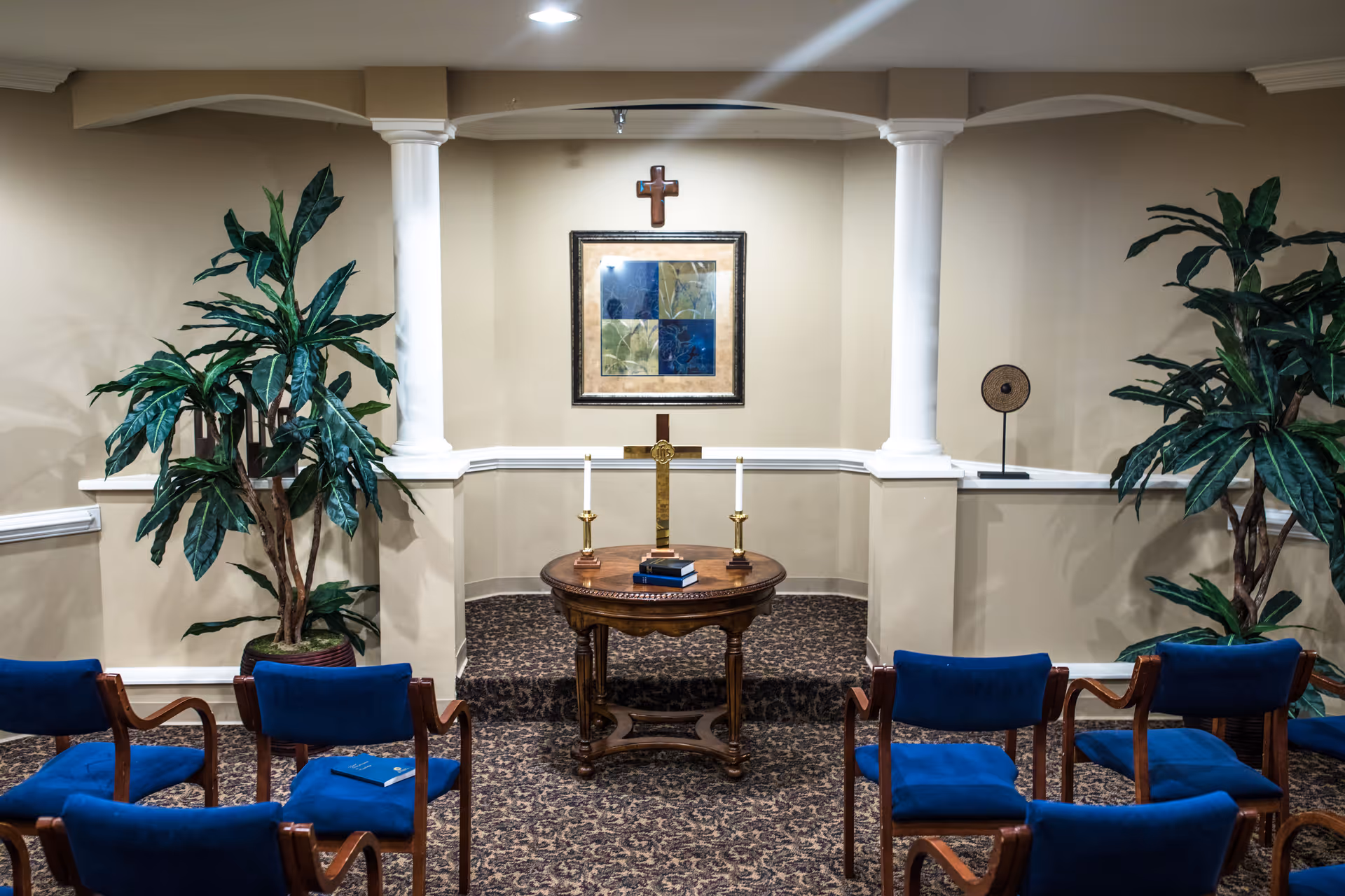 Small chapel-style interior with a wooden altar table, wall cross, candles, framed artwork, potted plants, and rows of blue chairs.