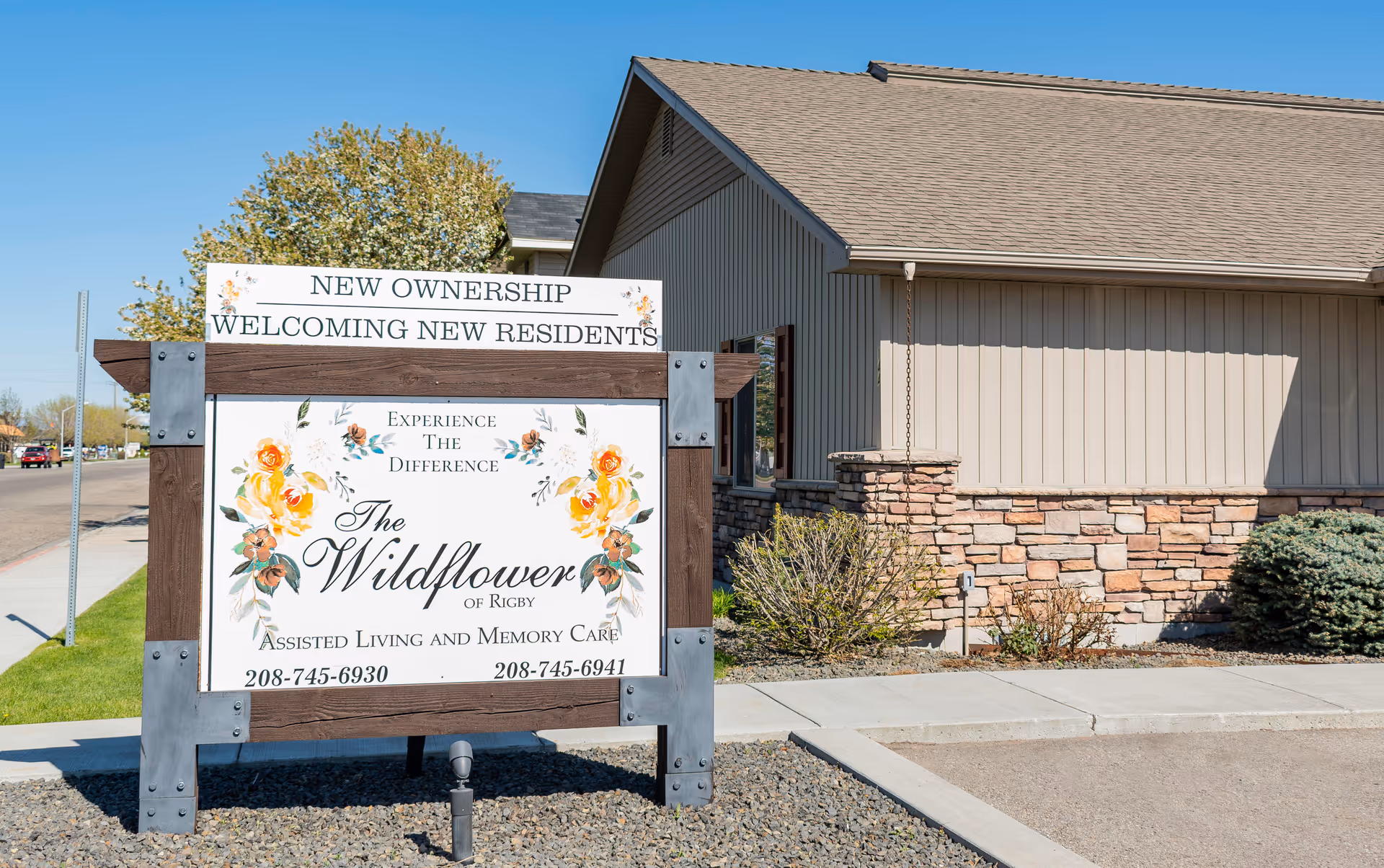Wooden sign welcoming new residents to The Wildflower of Rigby assisted living outside a beige building.