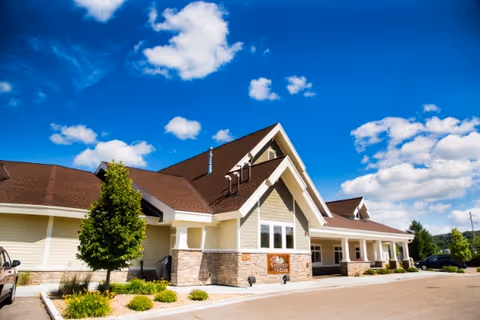 Exterior front of a single-story memory care facility with pitched roofs, stone and siding facade, landscaping and a driveway under a bright blue sky.