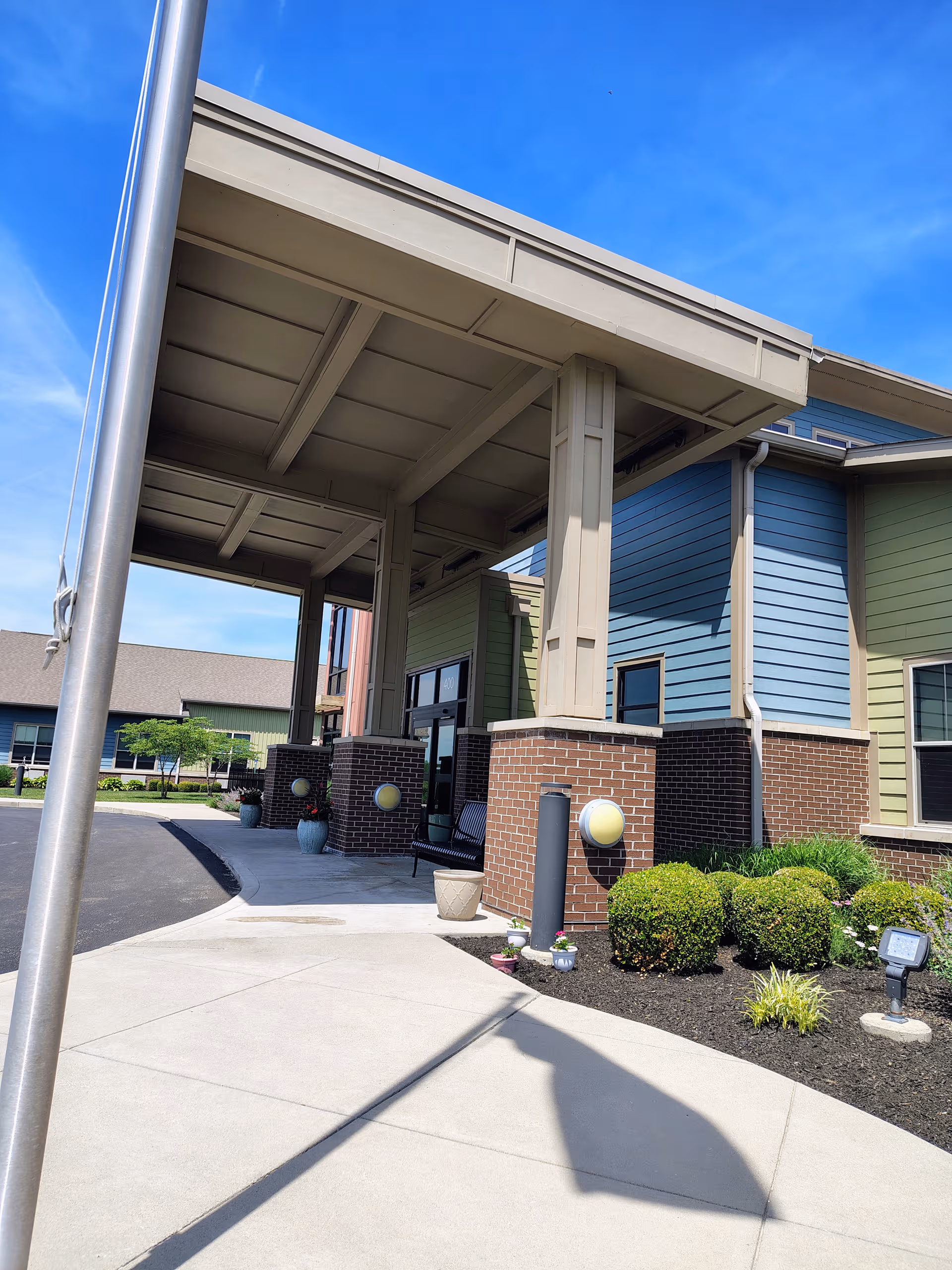 Entrance canopy over brick exterior with a bench, landscaped shrubs, and blue sky.