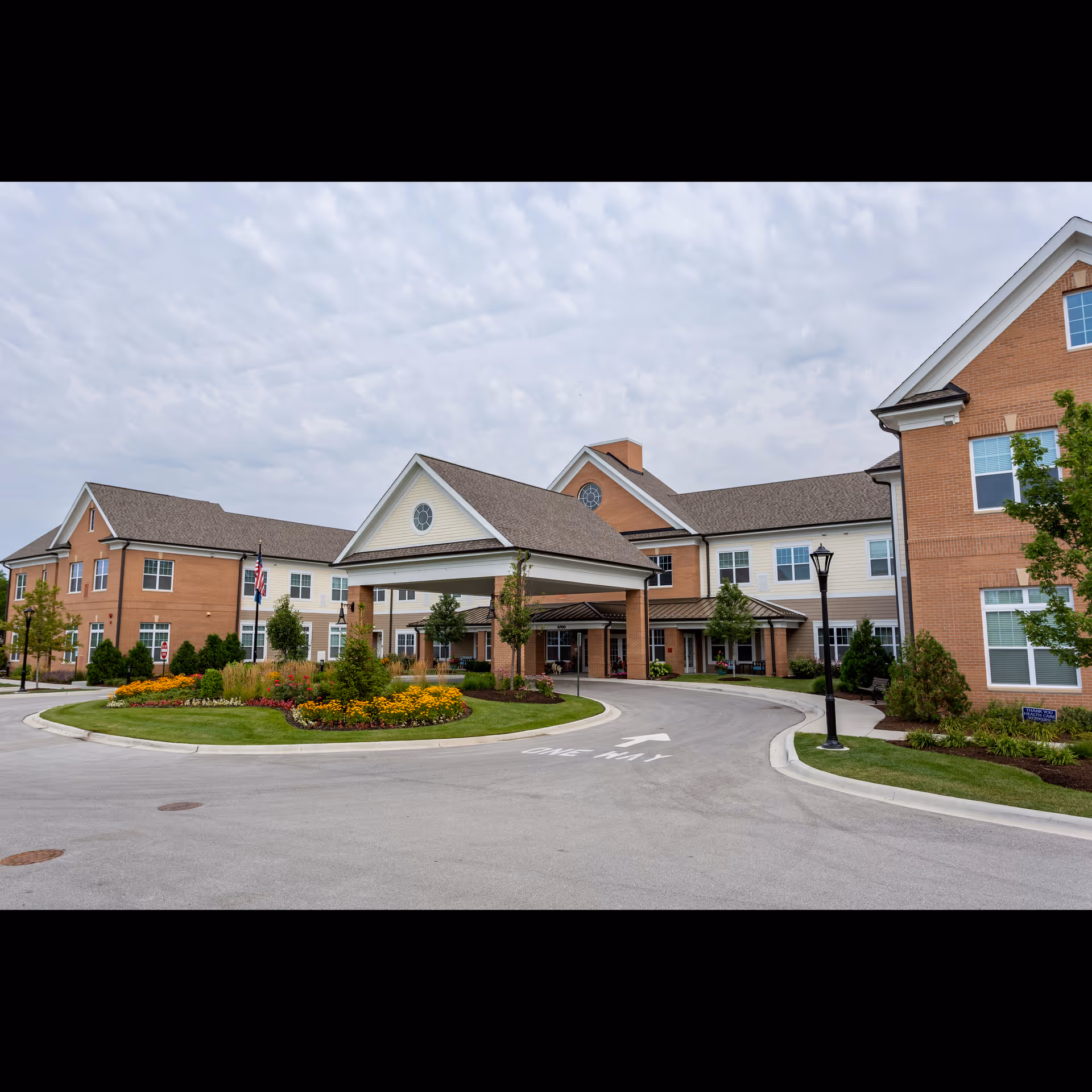 Exterior view of Arbor Terrace Glenview, a senior living facility with a large covered entrance, well-maintained landscaping including colorful flowers and green shrubs, and a circular driveway with a one-way sign painted on the pavement. The building features brick and light-colored siding with multiple windows and a peaked roof under a cloudy sky.