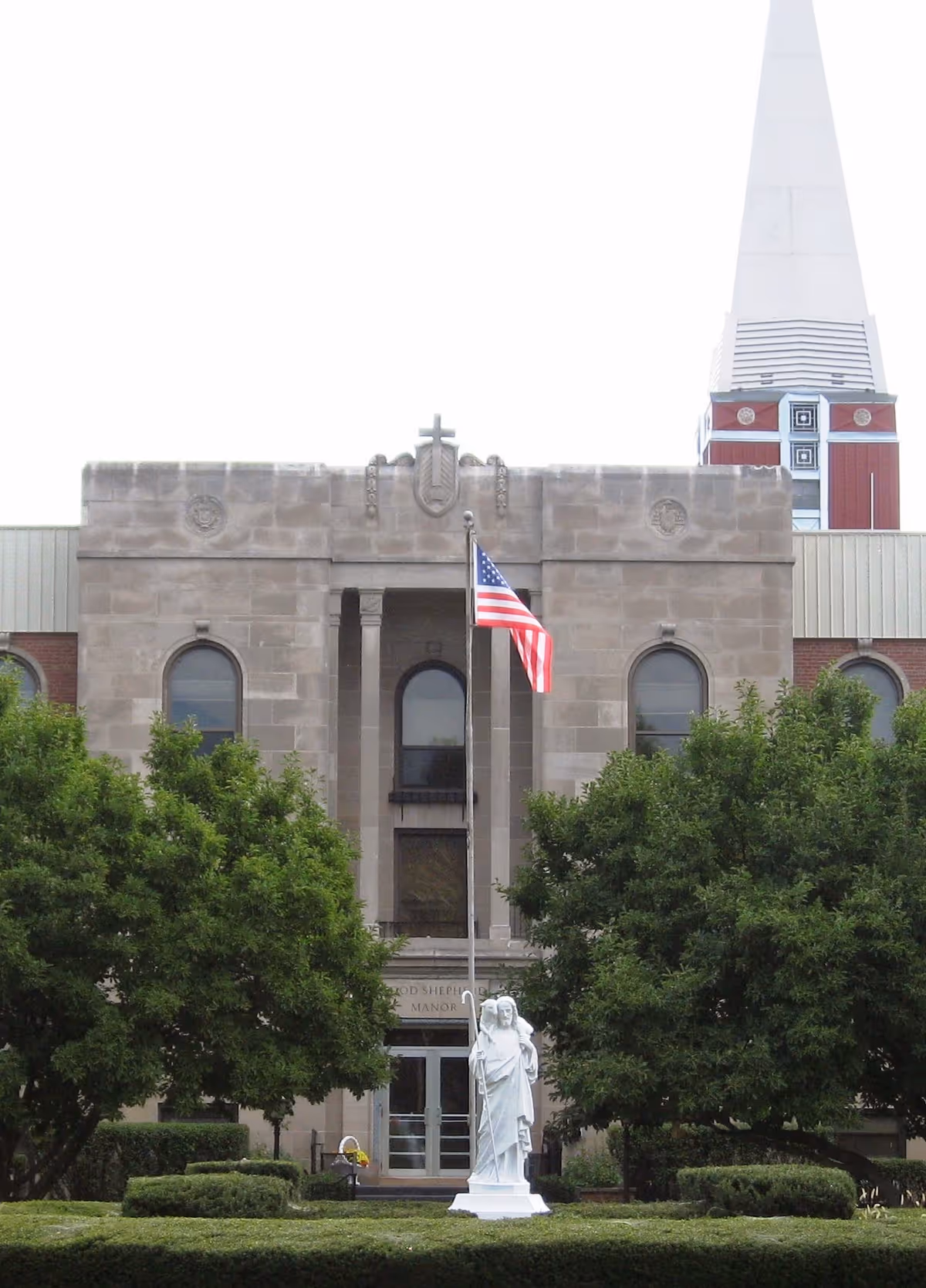Good Shepherd Manor building exterior with flag, statue, trees, and a church steeple.