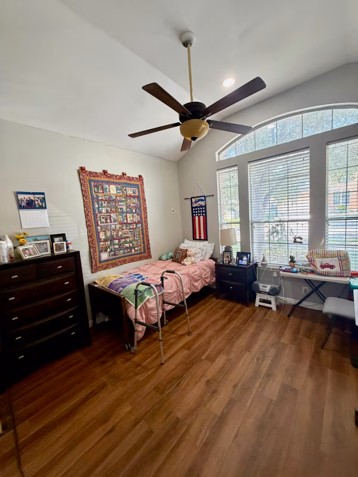 A bedroom in a care home with a single bed, dresser, walker, and large window.