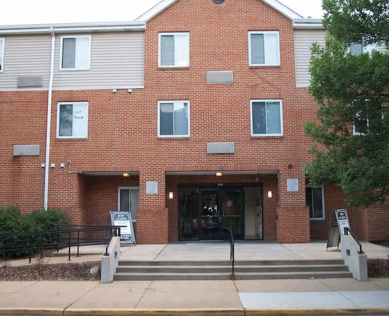 Brick three-story apartment building front with steps, a wheelchair ramp, and a glass double-door main entrance flanked by sandwich-board signs.
