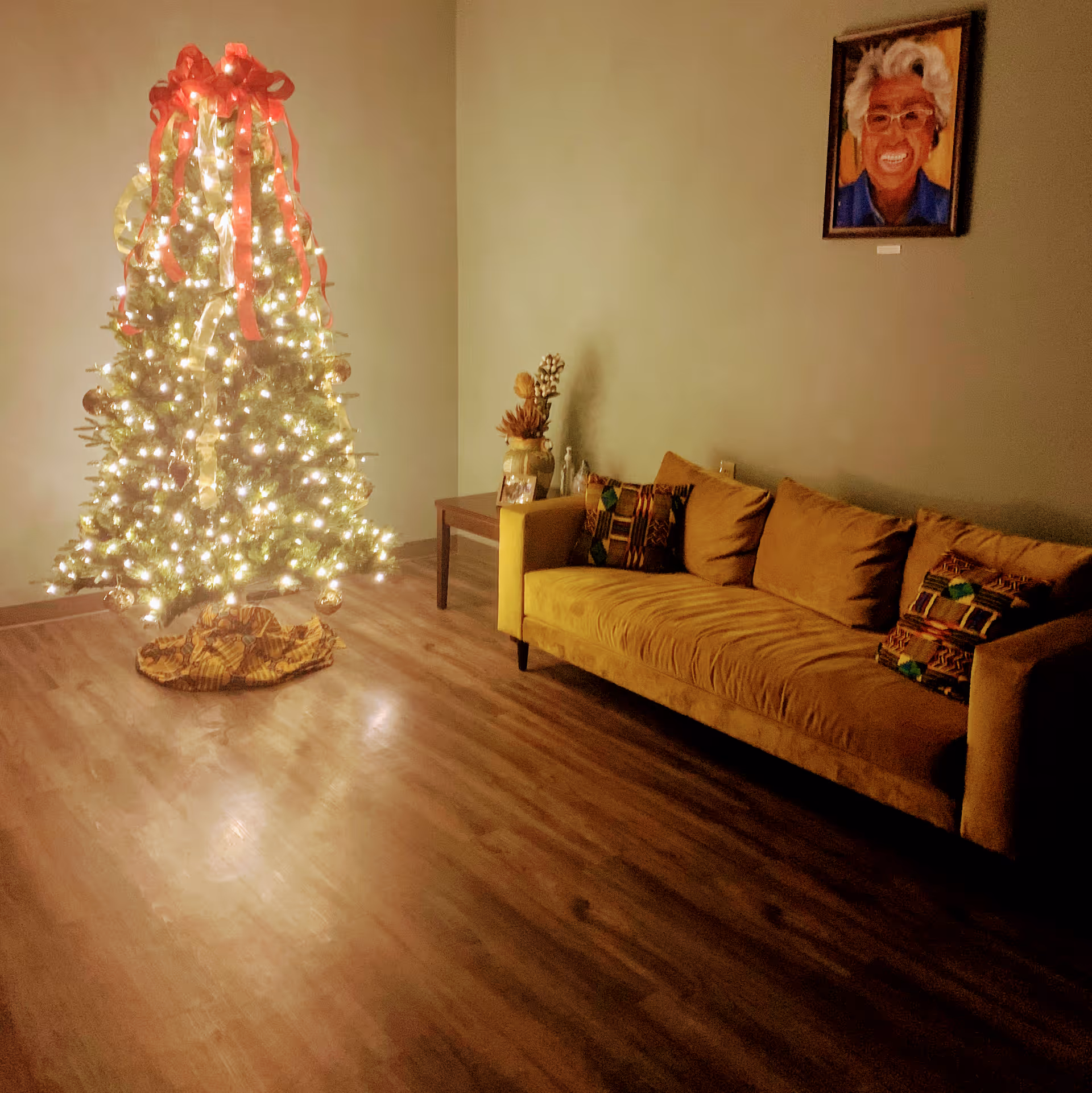 Warm living room with a lit Christmas tree beside a mustard sofa and beige walls.