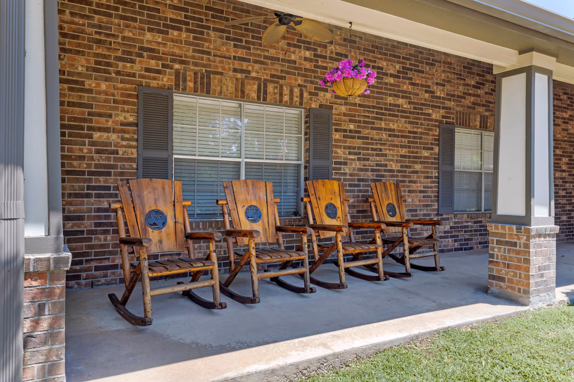 Front porch of brick senior living facility with five wooden rocking chairs and a hanging basket.