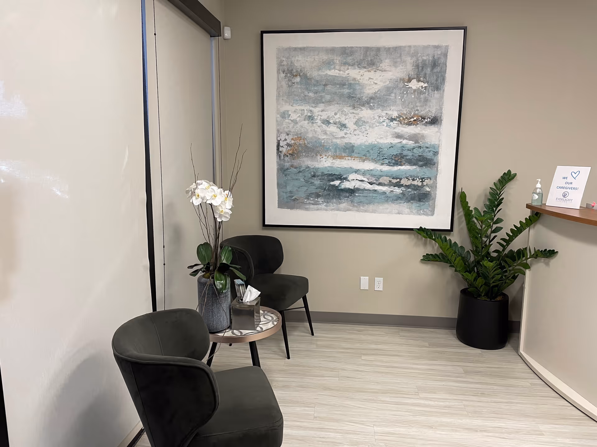 A waiting area with two dark chairs, abstract painting, plants, and a small table.