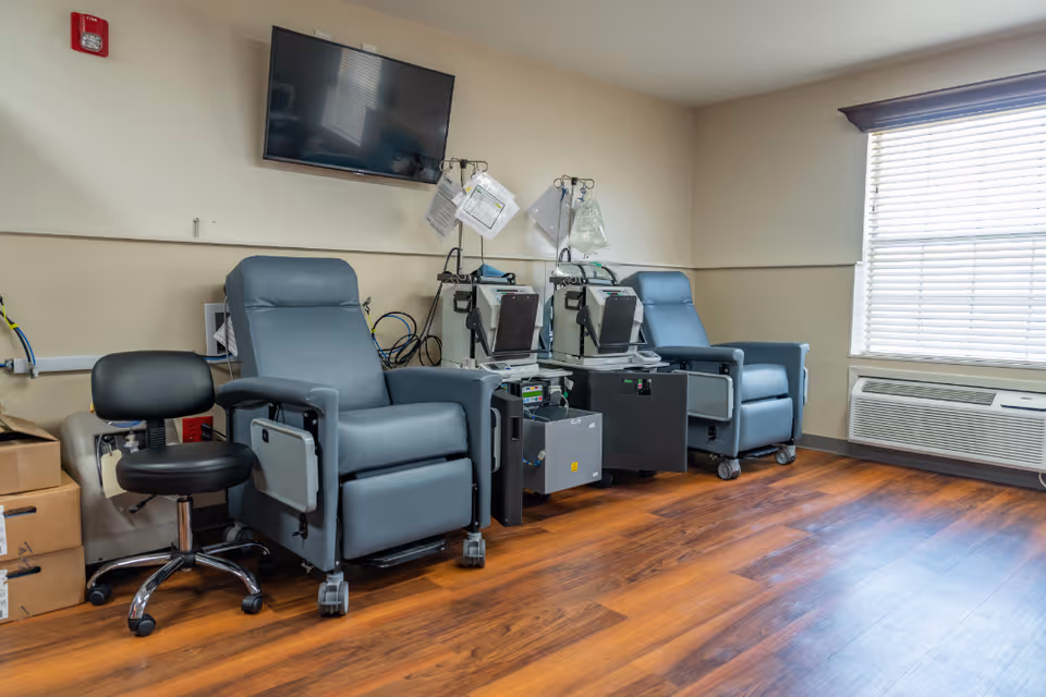 Medical treatment room with reclining dialysis chairs, dialysis machines, a wall-mounted TV and a window with blinds.