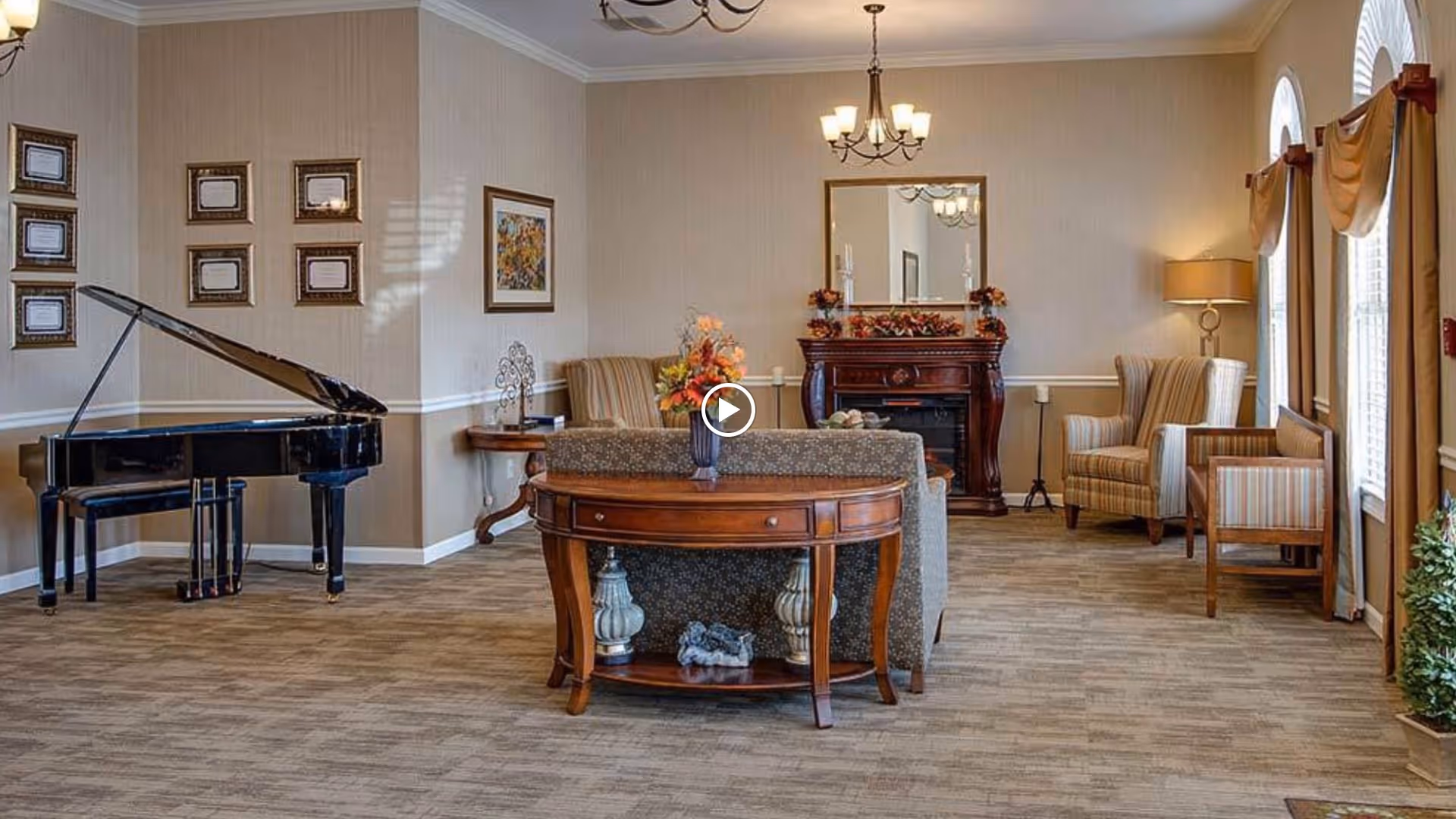 A cozy living room area with a grand piano on the left, a wooden console table with decorative items in the center, and comfortable armchairs near windows with curtains on the right. The room features a fireplace with a mirror above it, framed pictures on the walls, and warm lighting from a chandelier and a floor lamp.