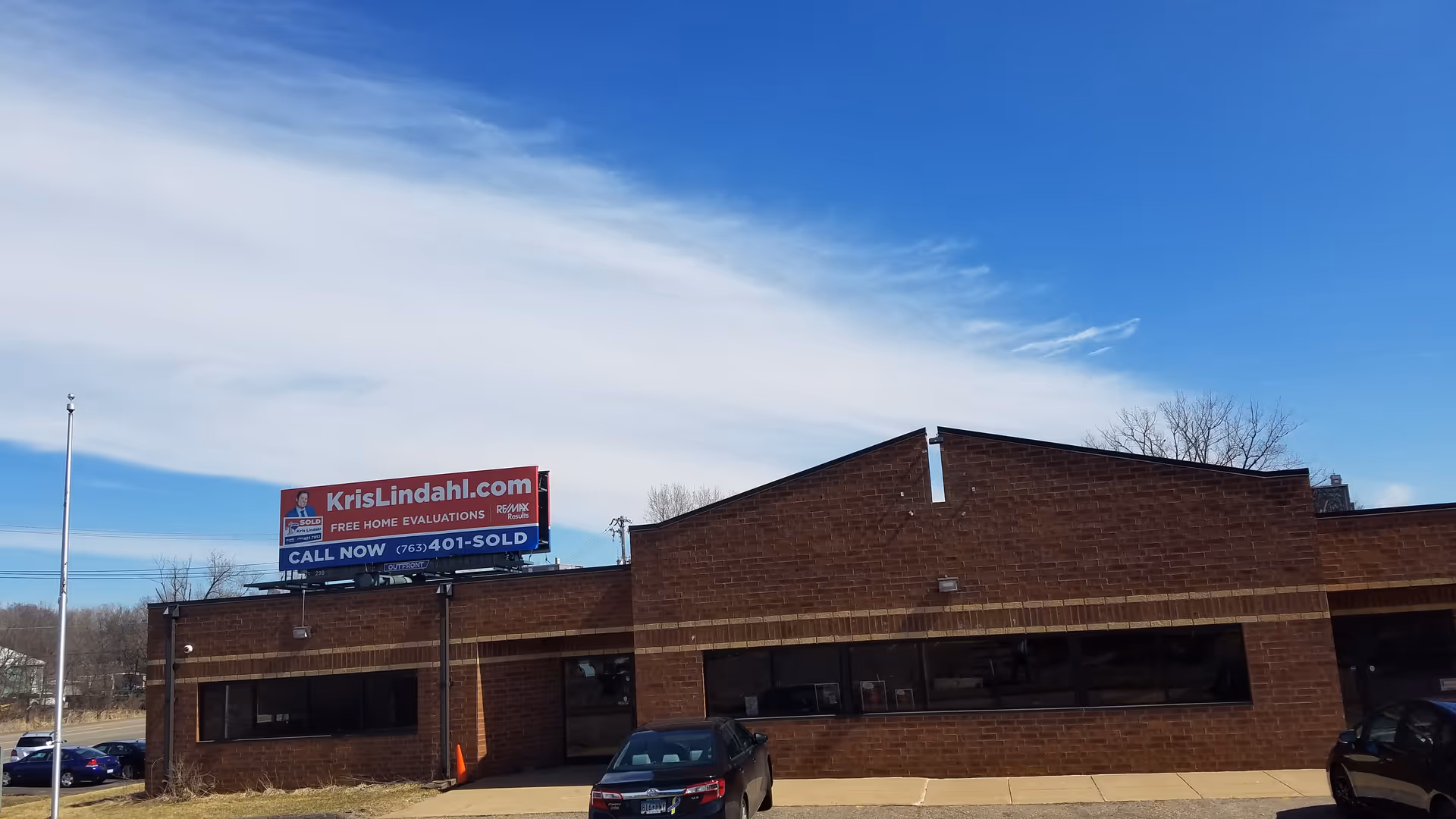 Brick building exterior with large windows and a Kris Lindahl billboard, with parked cars in front.