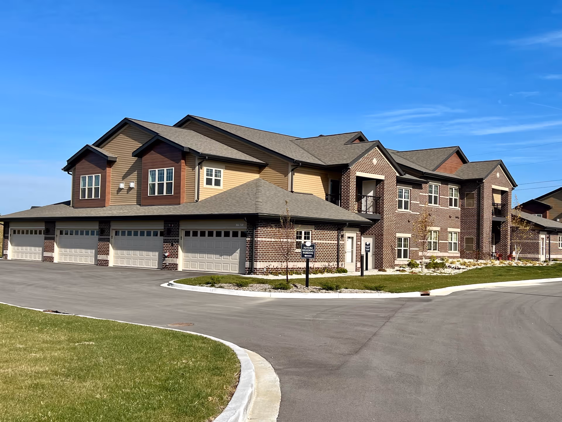 Exterior view of a two-story residential building with a combination of brick and siding facade under a clear blue sky. The building features multiple garage doors on the ground level and several windows on both floors. There is a paved driveway and a small landscaped area with grass and young trees in front.