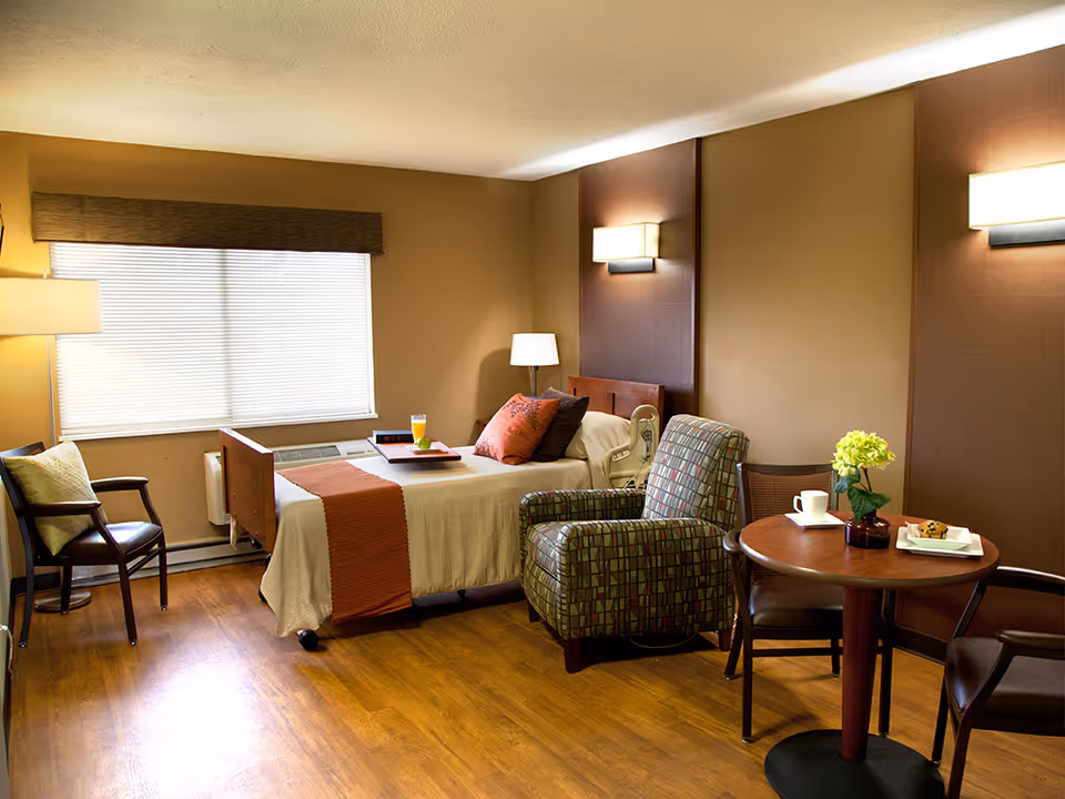 A cozy hospital-style bedroom with a bed, seating area, table, and warm lighting.