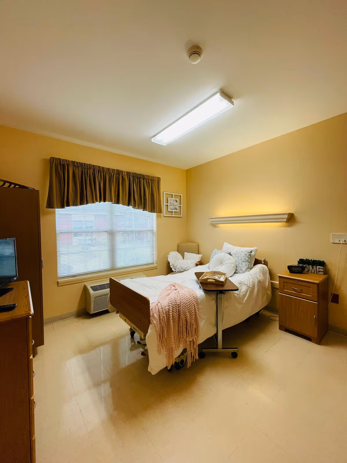 A small, neatly arranged bedroom in a healthcare facility with a single hospital-style bed covered with white bedding and decorative pillows. A pink throw blanket is draped over the foot of the bed. There is a wooden nightstand with a small plant and decorative items, a window with blinds and a valance, a wall-mounted light above the bed, and a wooden cabinet with a TV on top. The walls are painted a warm beige color.