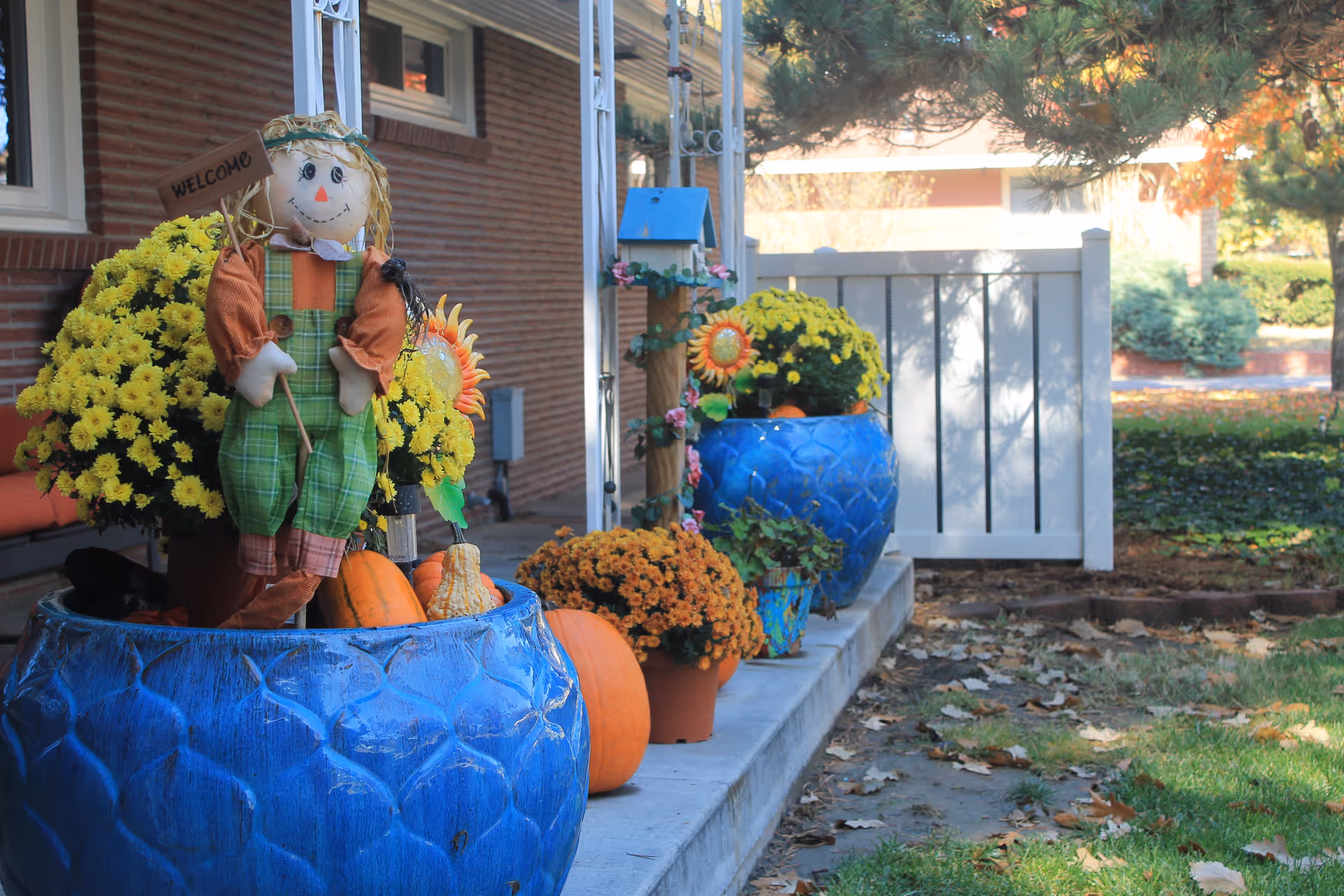 A front porch decorated with large blue pots filled with yellow and orange flowers, pumpkins, and a small scarecrow holding a welcome sign. The porch is next to a brick building with a white fence and trees in the background.