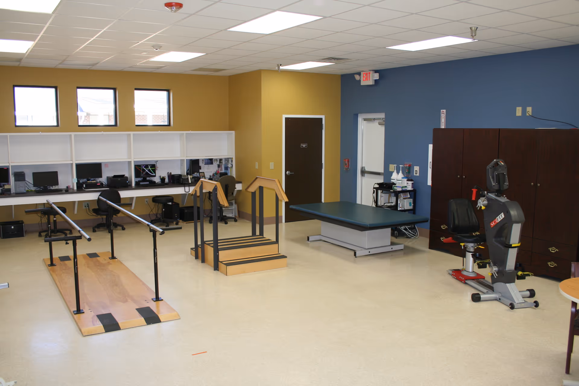 Rehabilitation gym with parallel bars, a treatment table, and exercise equipment in a bright room.