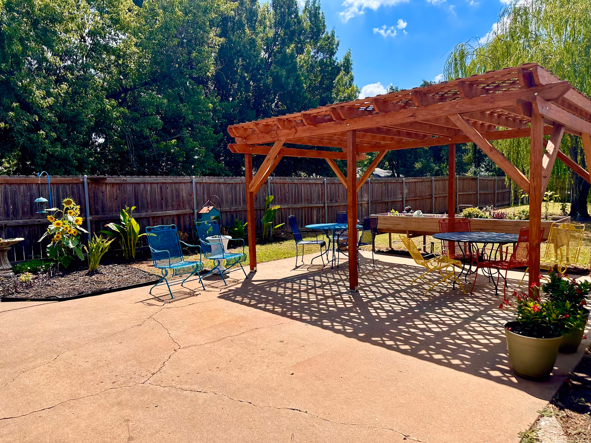 Sunlit backyard patio with a wooden pergola, colorful chairs, tables, potted plants, and sunflowers.