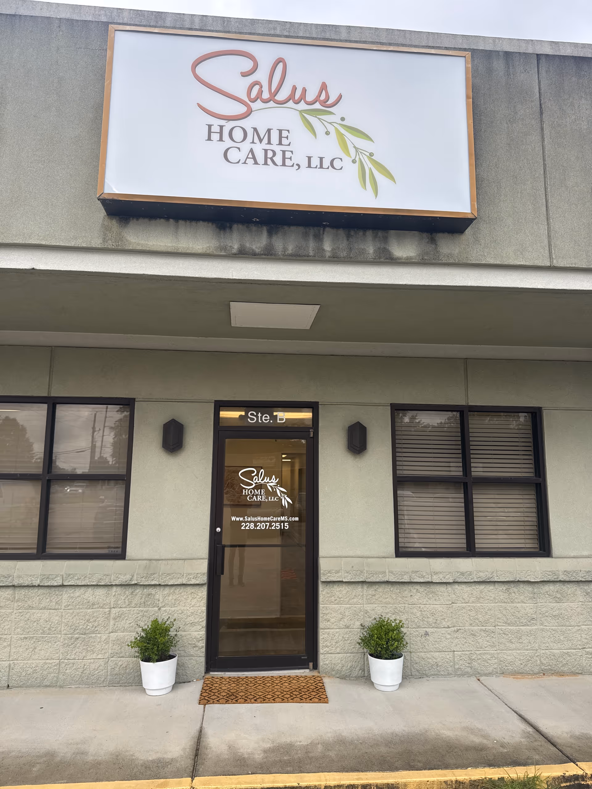 Exterior entrance of Salus Home Care, LLC, with sign, windows, and two potted plants.