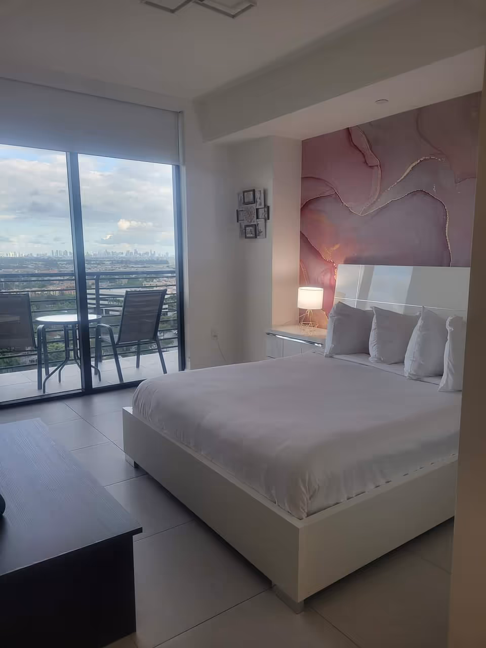 Modern bedroom with white bed, nightstand, and balcony doors revealing a city view.