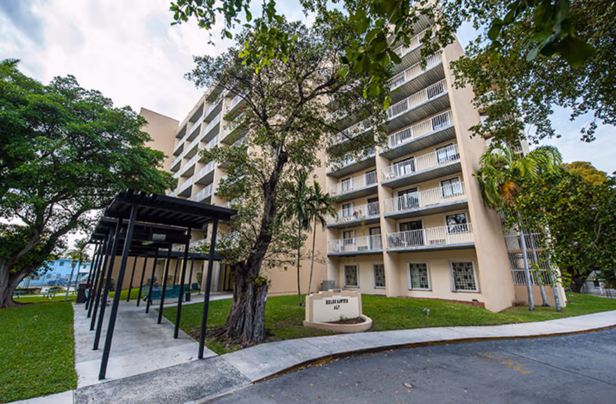 Front exterior of a multi-story assisted living building with balconies, a covered walkway, and surrounding trees.