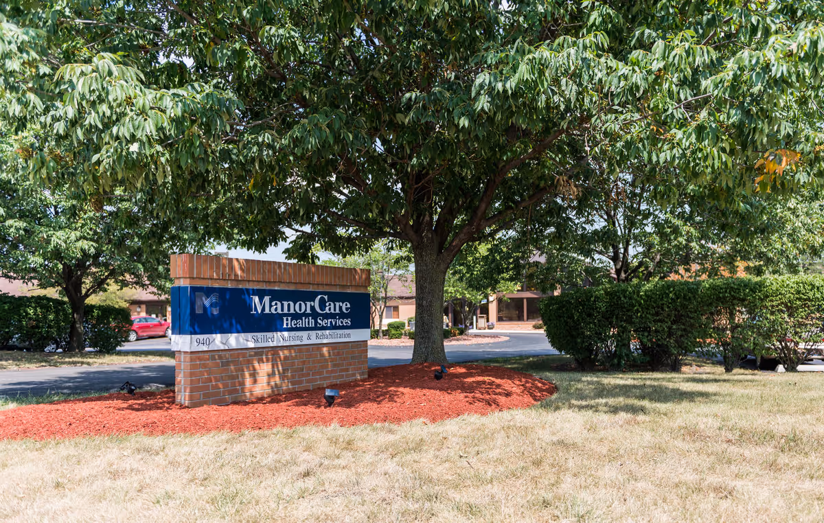 A brick ManorCare Health Services sign sits under a large tree on a manicured lawn.