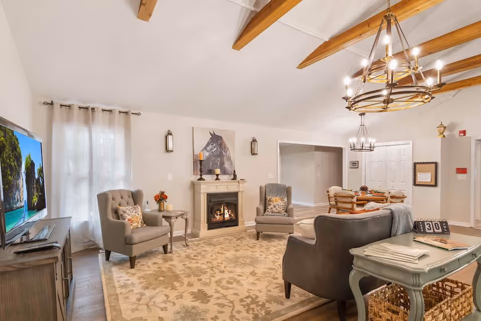 Spacious, warmly decorated living room with vaulted beamed ceiling, fireplace, armchairs, sofa, TV, and a small dining area.