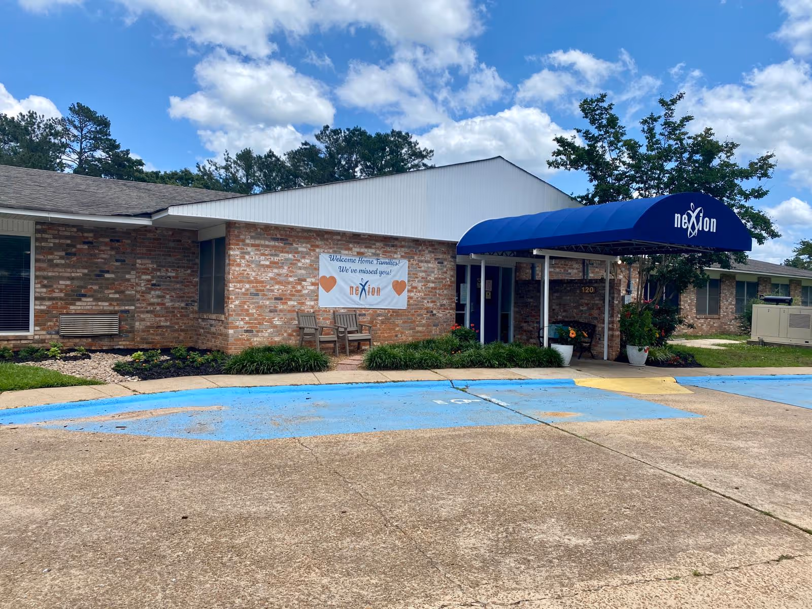 Front entrance of a single-story brick senior care building with a blue awning and a welcome banner.