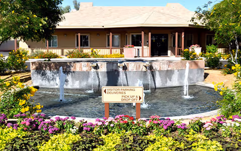 Front entrance of Paseo Village with a circular fountain, colorful flower beds, and a sign indicating visitor parking and drop-off.