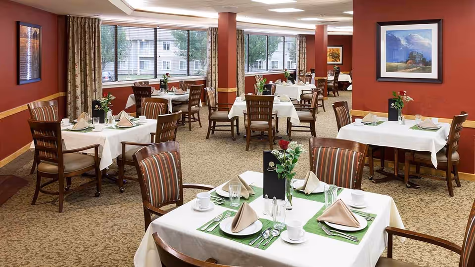 A dining room in a senior living facility with multiple tables set for meals. Each table is covered with a white tablecloth, green placemats, beige folded napkins, plates, cups, glasses, and silverware. There are wooden chairs with striped cushions around the tables. The room has red walls with framed artwork and large windows letting in natural light.