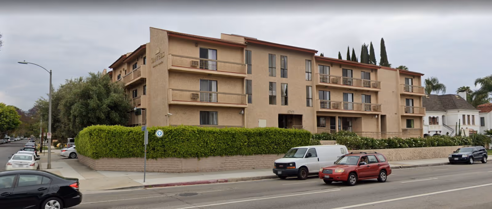 Exterior view of a three-story beige assisted living facility named Sunny Hills Assisted Living, surrounded by a green hedge and located on a street corner with parked cars and trees nearby.