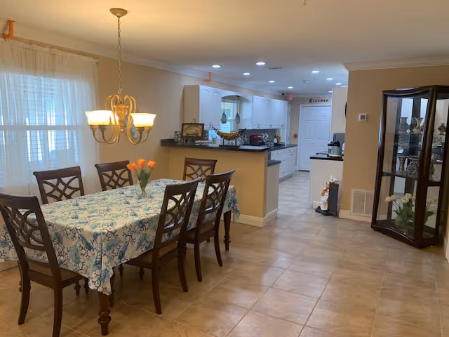 Bright dining area with a rectangular table covered by a blue floral tablecloth, six chairs, a chandelier overhead, and an open kitchen visible in the background.