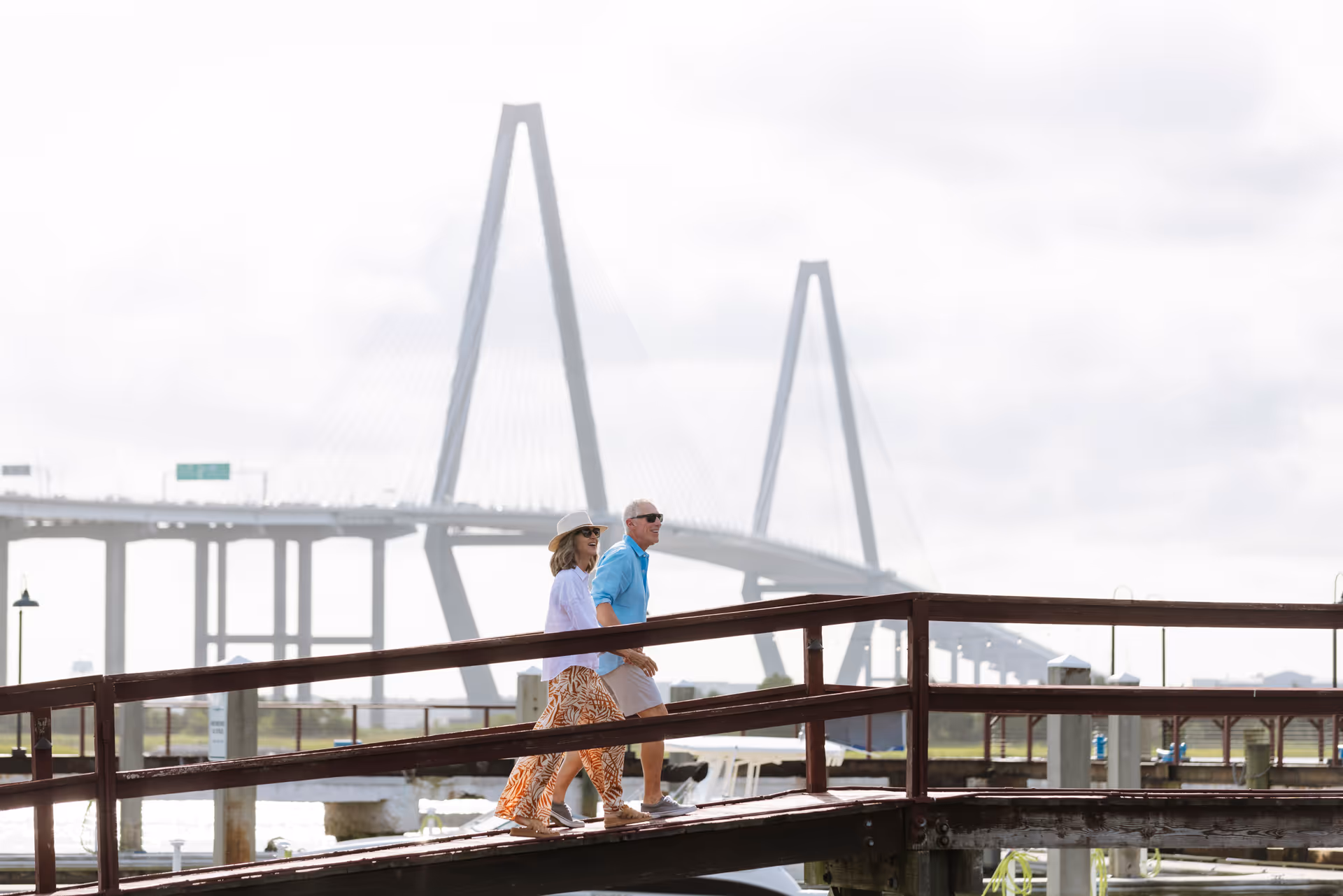 Two seniors walk along a wooden waterfront boardwalk with a large bridge in the background.