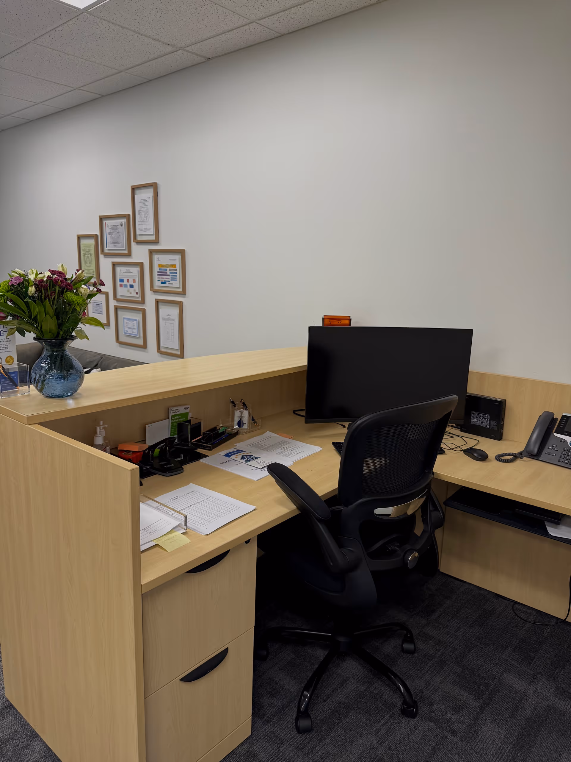 A wooden reception desk with computer monitor, chair, documents, and a vase of flowers in office.