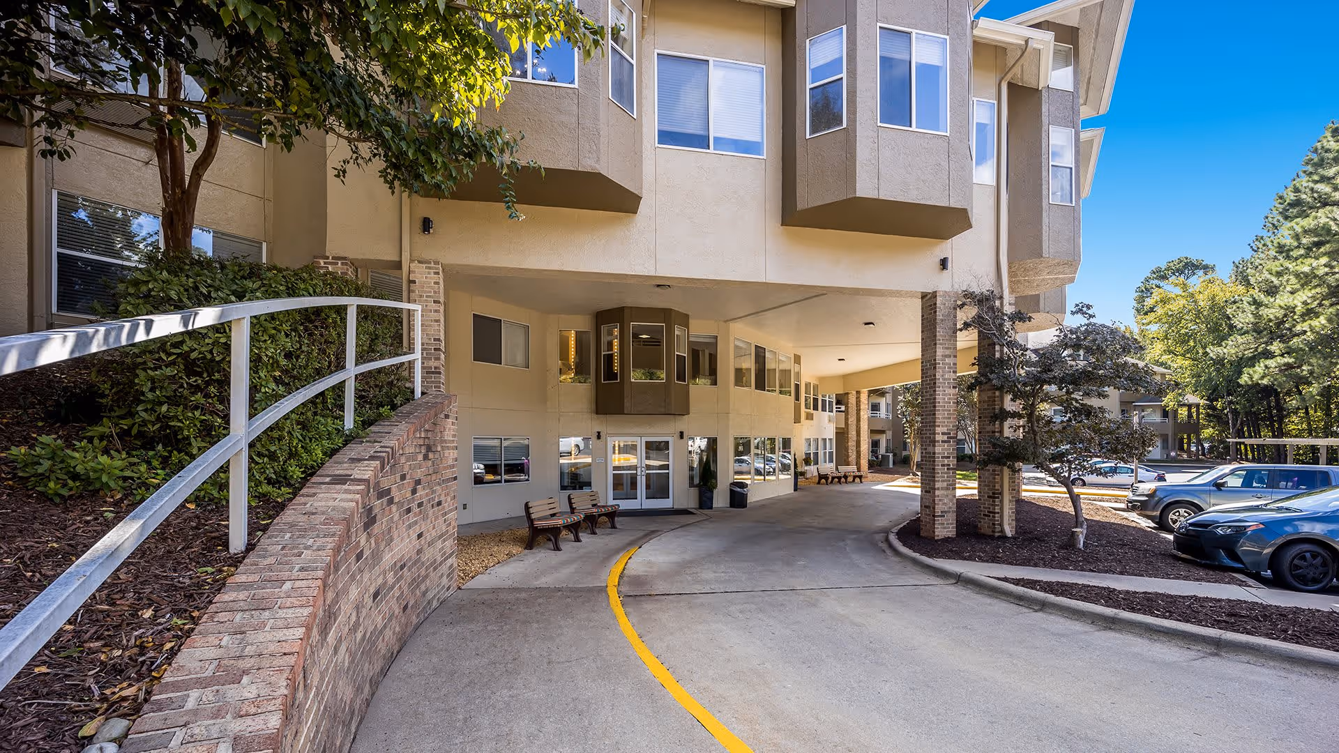 Entrance driveway of a senior living facility with a covered drop-off area, benches along the wall, and parked cars to the right. The building has beige walls with several windows and is surrounded by trees and landscaping.